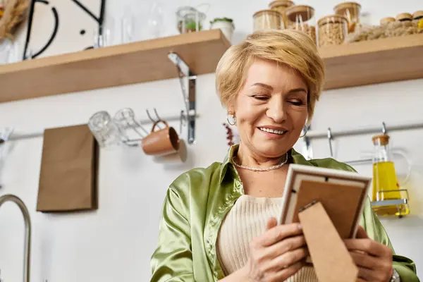 Mujer mayor con el pelo corto y rubio sonríe, sosteniendo una imagen enmarcada en su cocina brillante. - foto de stock