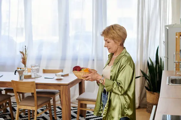A beautiful senior woman smiles while holding a basket of fresh fruits in a sunlit kitchen. — Stock Photo