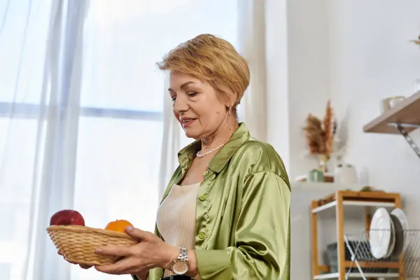 In a bright, warmly lit room, a beautiful senior woman fondly admires a basket of fresh fruits. — Stock Photo
