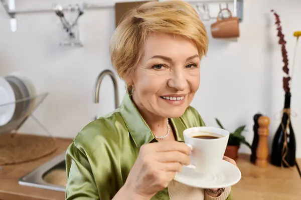 A beautiful senior woman holds a cup of coffee while smiling warmly in her bright kitchen. — Stock Photo