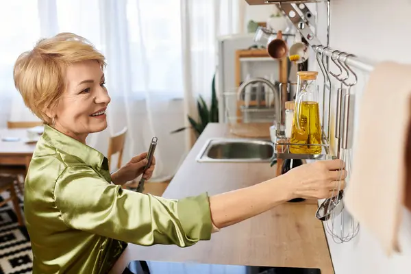 Senior woman smiles as she organizes utensils and prepares to cook in a sunny kitchen. — Stock Photo
