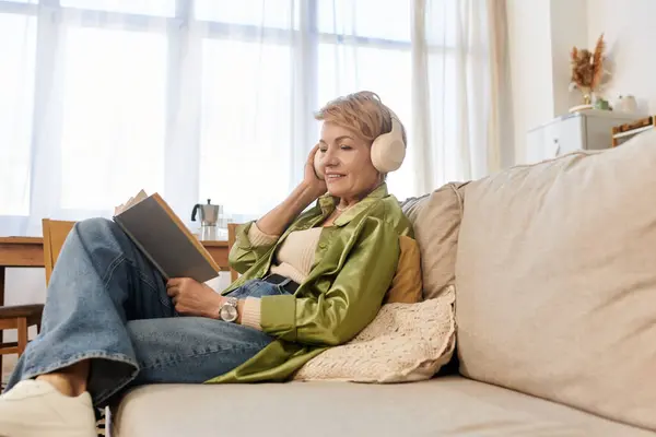 Mature woman relaxes on her sofa with headphones, reading a book in natural light. — Stock Photo