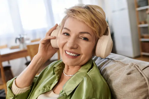 Mujer mayor con el pelo corto escucha música mientras se relaja en el sofá, irradiando felicidad. - foto de stock