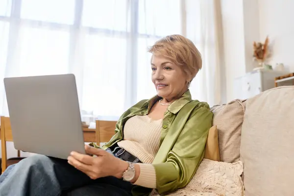 Senior woman with a warm smile engages with her laptop in a sunlit living area, showcasing joy. — Stock Photo
