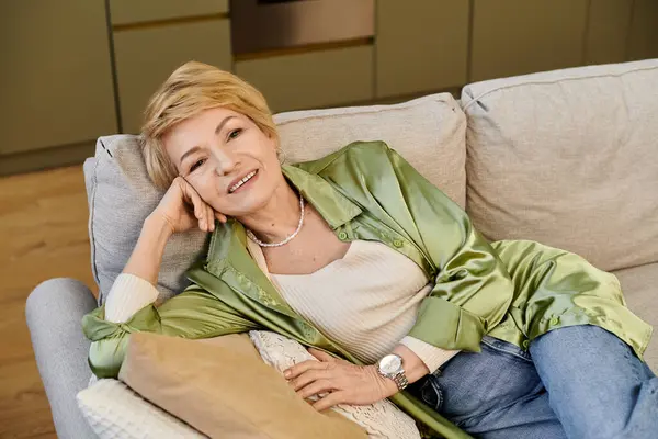 A beautiful senior woman relaxes elegantly on a cozy sofa, showcasing her graceful style and joy. — Stock Photo