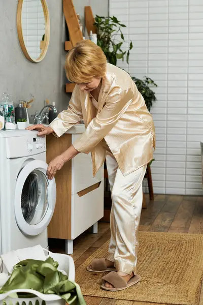 Eine schöne Seniorin in stylischer Loungewear bedient eine Waschmaschine und genießt dabei das häusliche Leben. — Stockfoto
