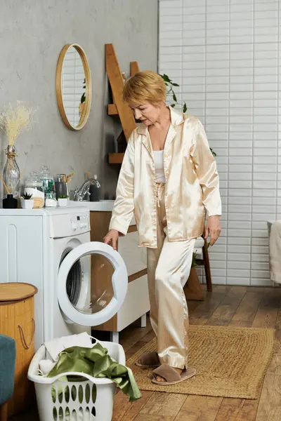 Eine hübsche Seniorin im seidenen Pyjama zieht zu Hause frische Wäsche aus der Waschmaschine. — Stockfoto