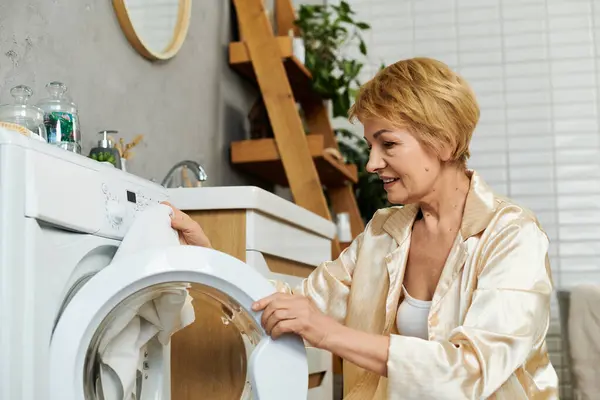 Glücklich reife Frau nimmt frisch gewaschene Kleidung aus der Maschine in einer heiteren Umgebung. — Stockfoto