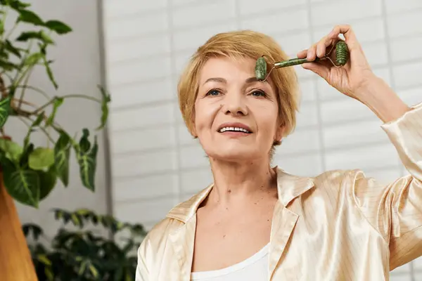 A happy senior woman applies a roller to her face in a bright, green filled room. — Stock Photo