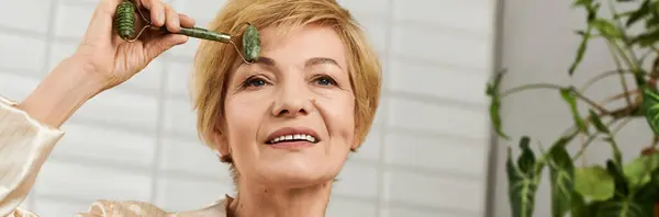Mature woman applies a jade roller to her face, embracing self care and relaxation at home. — Stock Photo