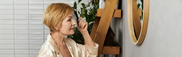 Una donna anziana in vestaglia di seta si pettina i capelli con uno specchio circondato da piante. — Foto stock