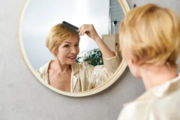 Un'elegante donna anziana sorride al suo riflesso mentre pettina i suoi capelli corti biondi. — Foto stock