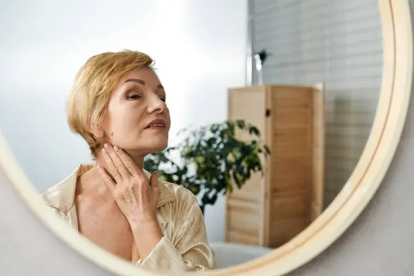 Una bella donna anziana si tocca delicatamente il collo mentre si guarda allo specchio, irradiando fiducia. — Foto stock