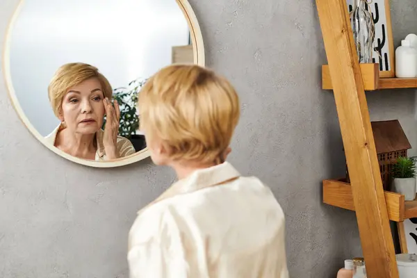 In uno spazio interno tranquillo, una bella donna anziana applica delicatamente la cura della pelle mentre si guarda in uno specchio. — Foto stock