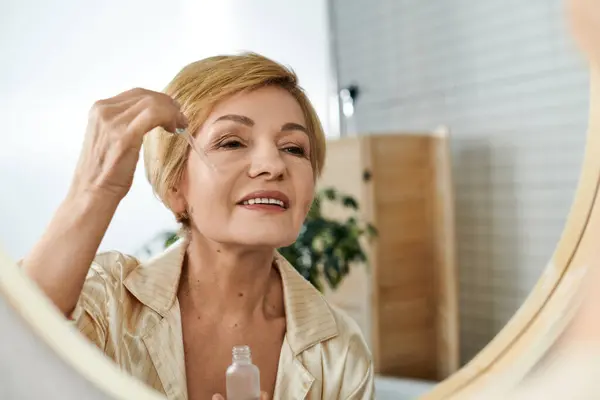 Donna matura sorride mentre applica il siero per la cura della pelle, circondata da luce naturale e decorazioni calmanti — Foto stock