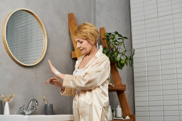 Una bella donna anziana applica la cura della pelle in un moderno bagno pieno di piante, sorridendo dolcemente.. — Foto stock