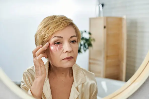 Una donna matura applica macchie agli occhi in un bagno calmo, dando priorità alla sua routine di cura della pelle. — Foto stock
