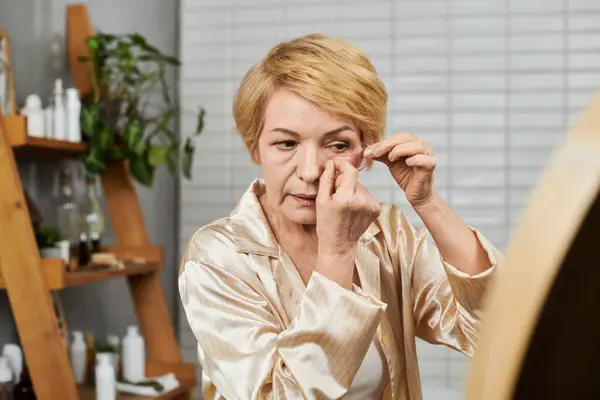Una bella donna anziana applica con attenzione la cura della pelle in un bagno tranquillo pieno di verde. — Foto stock