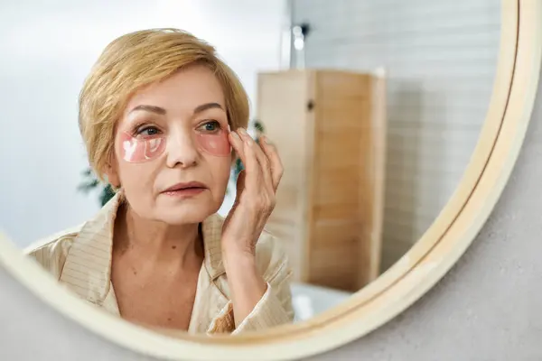 Donna matura si applica sotto le macchie sugli occhi di fronte a uno specchio rotondo durante la sua routine di auto-cura. — Foto stock
