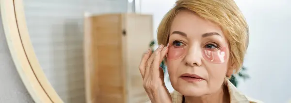 Beautiful mature woman applies soothing patches under her eyes, enhancing her skincare routine. — Stock Photo