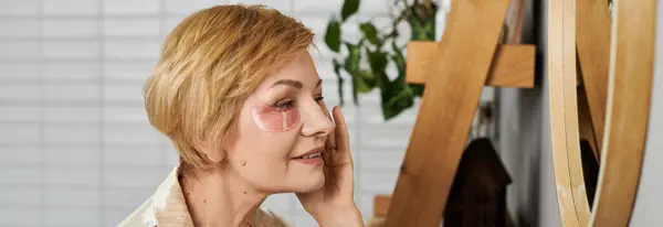 Bella donna matura che sorride delicatamente mentre applica patch per la cura della pelle nel suo luminoso spazio vitale — Foto stock
