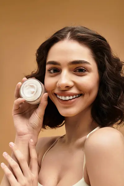 Hübsche junge Frau unterstreicht ihre Hautpflege-Routine mit einem Glas Creme und lächelt selbstbewusst. — Stockfoto