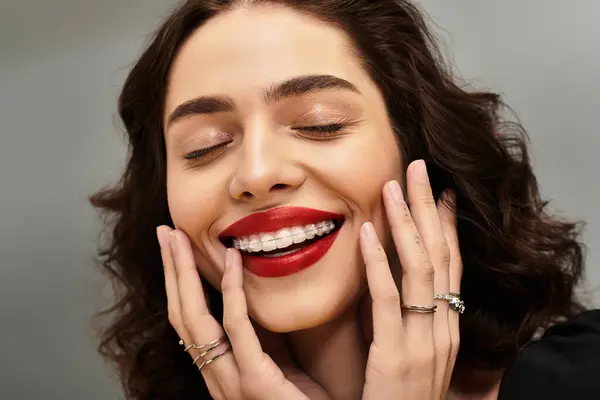 A young woman is smiling with her hands on her cheeks, showcasing her braces and bold red lips. — Stock Photo