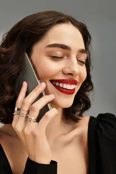 Uma jovem mulher com aparelho sorri brilhantemente em seu smartphone contra um fundo cinza. — Fotografia de Stock