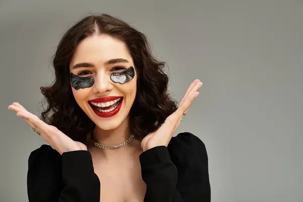 Una giovane donna radiosa mette in mostra i suoi bretelle e labbra rosso brillante, trasudando fascino su uno sfondo grigio. — Foto stock