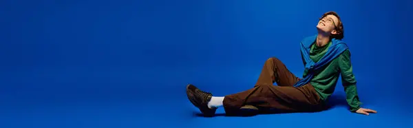 A young man leans back against a blue background, displaying stylish attire and a chill vibe. — Stock Photo