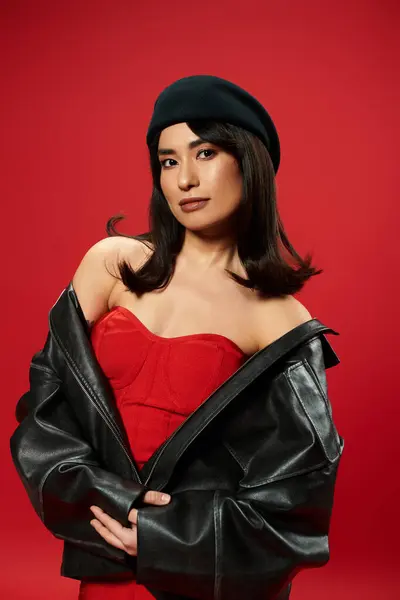 A young Asian woman flaunts a chic red outfit and black leather jacket against a bold red backdrop. — Stock Photo