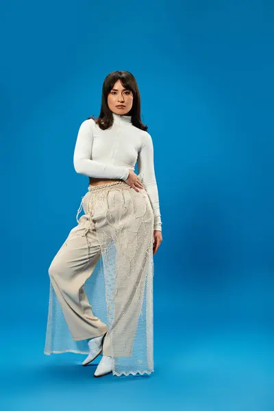 A stylish young Asian woman confidently poses in trendy attire against a striking blue backdrop. — Stock Photo