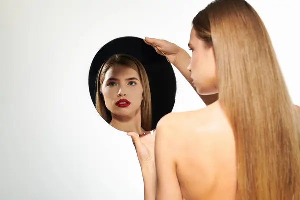 Una giovane donna elegante con i capelli lunghi ammira il suo riflesso in uno specchio rotondo. — Foto stock