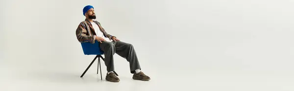 A stylish young man with a beard sits in a modern chair, flaunting his unique fashion. — Stock Photo