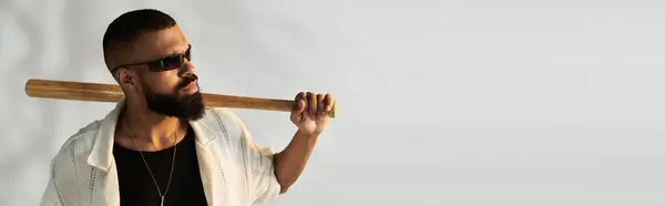 Un uomo barbuto in occhiali da sole posa con fiducia con un pipistrello sulla spalla in un ambiente moderno. — Foto stock