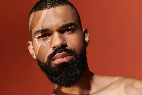 Handsome young man confidently posing with a striking facial design and warm reddish backdrop. — Stock Photo