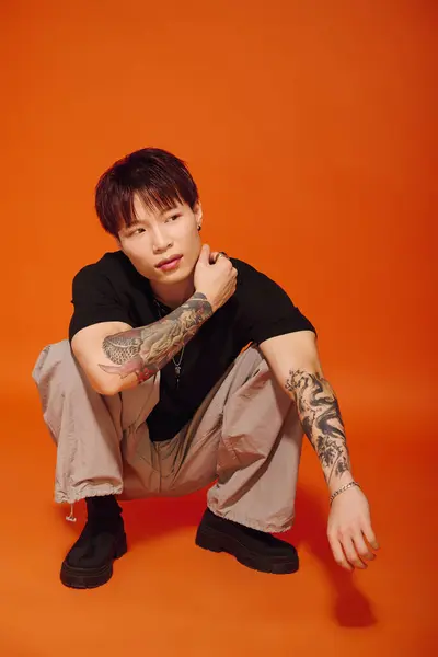 A handsome young Asian man crouches stylishly, showcasing his tattoos against an orange background. — Stock Photo