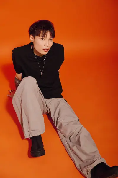 Уверенный молодой азиат в модном наряде сидит на полу на ярком оранжевом фоне. — стоковое фото
