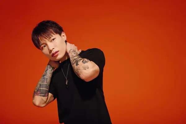 A stylish young Asian man poses confidently against a vibrant orange backdrop, showing off tattoos. — Stock Photo