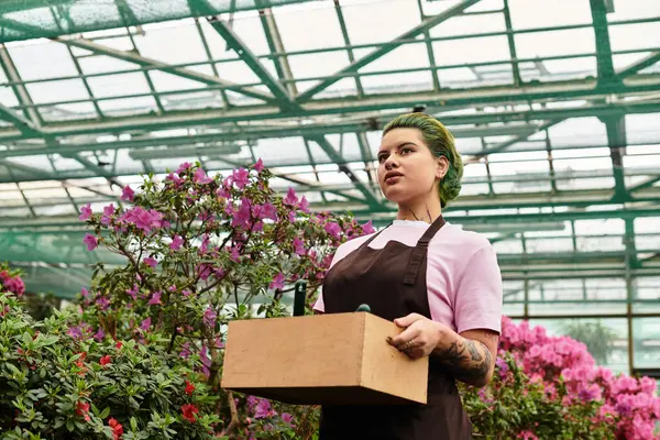 A young woman joyfully works in a blooming greenhouse, holding a wooden box of gardening tools. — Stock Photo