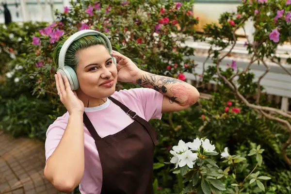Amidst blooming flowers, a young woman in a greenhouse listens to music with joy while working. — Stock Photo