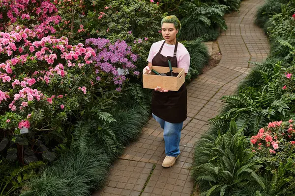 In a lively greenhouse, a young woman carries a wooden crate while surrounded by colorful blooms. — Stock Photo