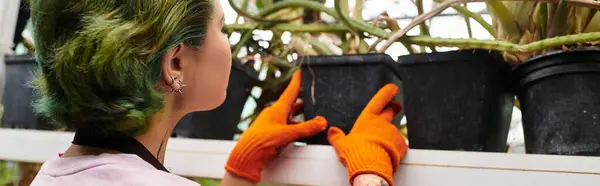 Young female gardener with green hair works diligently among potted plants in a greenhouse. — Stock Photo