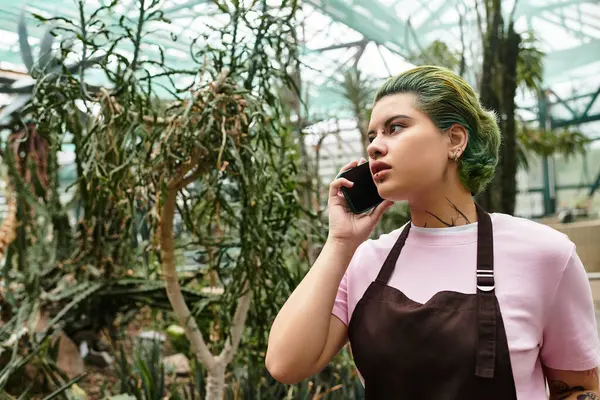 A young woman with green hair talks on the phone amidst lush greenery in a greenhouse. — Stock Photo
