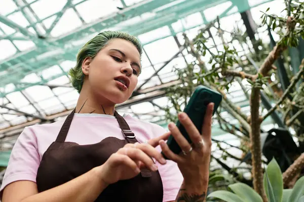 Una joven con una camisa rosa y un delantal comprueba su teléfono mientras cuida plantas en un invernadero. - foto de stock