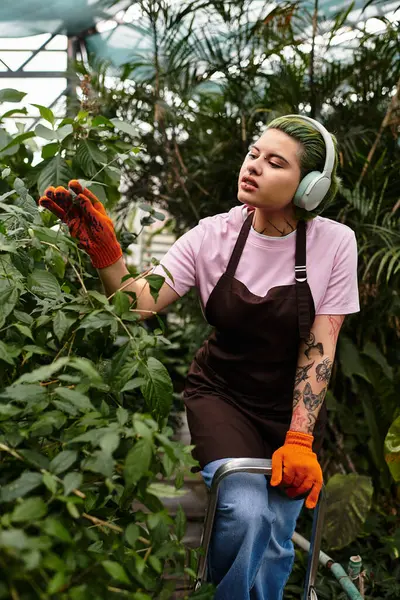 Amid lush greenery, a young woman cares for plants in a colorful greenhouse while enjoying music. — Stock Photo