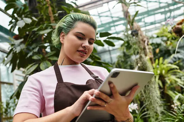 In einem üppigen Gewächshaus navigiert eine junge Frau gekonnt ihr Tablet, während sie sich mit Pflanzenpflege beschäftigt. — Stockfoto