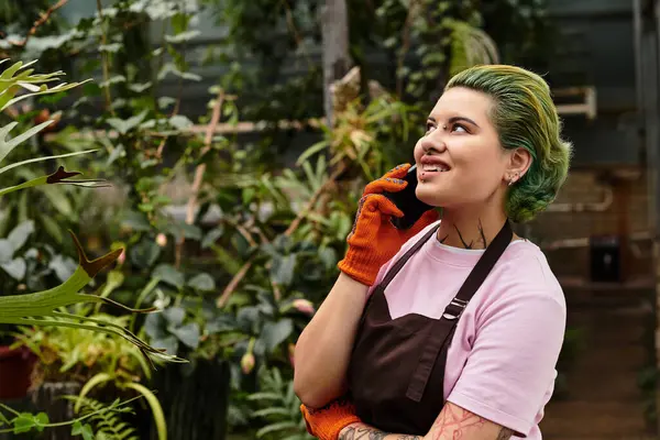 Young woman in a vibrant greenhouse talks on her phone while surrounded by flourishing plants. — Stock Photo