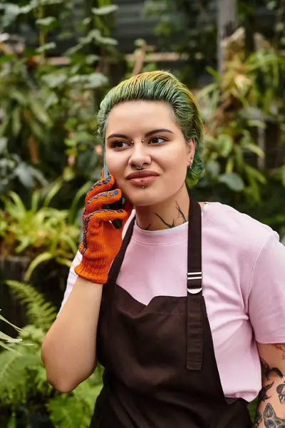 In a vibrant greenhouse, a young woman smiles on the phone, wearing gardening gloves. — Stock Photo