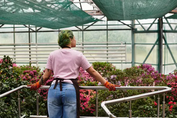 A young woman with green hair admires blooming flowers while working in a serene green house. — Stock Photo